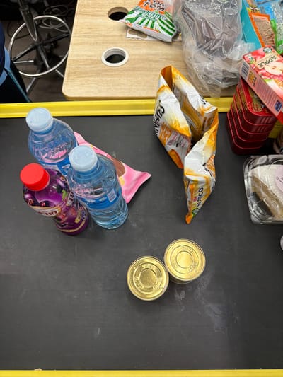 The scene depicts a collection of consumer goods arranged on a dark grey retail counter with a yellow strip along its edge, located within a commercial establishment in Babban Giji, Nigeria.

In the immediate foreground on the dark grey counter, two clear plastic bottles of water with light blue caps are present. Adjacent to these is a purple-labeled bottle with a red cap, identified as "RIBITO Blackcurrant" flavored drink. Partially obscured behind these bottles is a pink rectangular packet, possibly containing snacks. To the right, two identical gold-colored cylindrical containers with concentric ring patterns on their tops are positioned side by side.

Further back on the counter, a partially opened or consumed bag of "NASCO Pancakes Original" is visible, its white and orange packaging crumpled.

The background transitions to a light brown wooden surface, possibly an adjacent shelf or workstation, featuring two circular cutouts, one containing a white plastic grommet. Scattered across this wooden surface are several items: a green and white striped wrapper (possibly "Tom Tom" brand candy), various other items wrapped in clear plastic, a stack of four red square-shaped containers, and a white and red box labeled "APPLE". A clear plastic container holding a piece of white-topped dessert, likely a cake, is also visible. To the far left, the bottom section of a metal chair base with wheels is partially visible, alongside a small portion of a blue garment. No individuals are visible in the frame, but the arrangement of items suggests a retail transaction or preparation area.