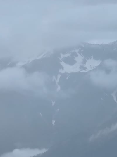 An outdoor, elevated medium shot captures a mountain range shrouded in dense, grey clouds and mist. The peaks of the mountains are partially visible, with patches of snow scattered across their surfaces. The lower slopes are obscured by a thick layer of fog, with only faint outlines of trees visible at the very bottom of the frame. The overall atmosphere is somber and ethereal, conveying a sense of remoteness and the dramatic power of nature. The scene is consistent with the natural landscape surrounding Prini, India, a town nestled in the Himalayas known for its mountainous terrain and unpredictable weather.