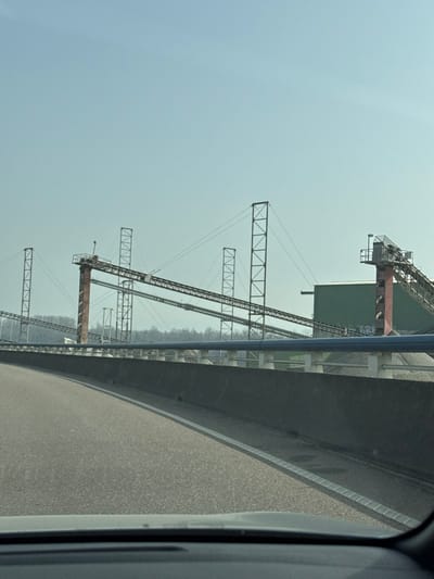 The image depicts an industrial material handling facility, located in Altenheim, Germany, observed from a vehicle's interior. In the foreground, the top edge of a vehicle dashboard is visible, along with a section of an asphalt road featuring a solid white edge line. A dark grey concrete barrier separates the road from an area containing piles of light-colored aggregate. Above this barrier, a series of light blue pipes are supported horizontally by white pedestals. The middle and background are dominated by an extensive network of elevated conveyor belt systems. These systems are supported by numerous tall, dark-colored lattice towers and substantial reddish-brown cylindrical structures. A large, dark green corrugated metal building is visible on the right, integrated into the conveyor infrastructure. Light-colored graffiti is present on a lower support section adjacent to the green building. A distant horizon line of leafless trees is visible beneath a clear, light blue sky. No individuals or active operations are discernible within the frame.