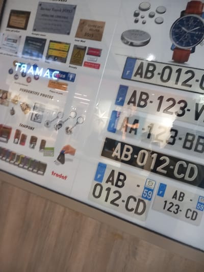 This image displays a brightly lit commercial display case or counter within a store located in Vendin-le-Vieil, France. The scene is dominated by a variety of customizable products and informational signage, presented on a white background with a light-toned wooden floor visible beneath.
On the upper left, multiple rectangular plaques are arranged. One prominently features "Docteur Franck DUPAS, MÉDECINE GÉNÉRALE," while another indicates "CUIVRE DES GUEULES 2019." Other plaques contain text and warning symbols, including a yellow triangle with an exclamation mark. Below these, a blue neon-style sign is visible, displaying "CAMART" (reflected as "TRAMAC"), with a smaller sign beneath reading "PENDENTIFS PHOTOS."
The lower left section showcases an array of small metal objects, including various keychains and pendants. Adjacent to these, a line of self-inking stamps in different colors is displayed, identifiable by the "trodat" brand. An interaction is depicted where a disembodied hand presses one of these stamps onto a white surface.
The upper right portion of the display features several circular metallic objects, resembling coins or tokens, of varying sizes. A wristwatch with a blue face and a brown leather strap is also prominently displayed.
The lower right section presents multiple sample French vehicle license plates. Examples include white plates with black lettering such as "F AB-012-CD," "F AB-123-V," and "F AA-123-BB," many featuring the European 'F' insignia and the regional department number '59'. A black license plate with white lettering, displaying "AB-012-CD," is also present.
The entire display surface exhibits reflections, including fragmented images of the surroundings and possibly the person capturing the image.