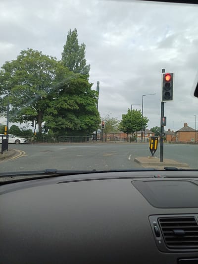 The image depicts a street scene in Middlesbrough, United Kingdom, from the perspective of a driver. The sky is overcast, and the scene is dominated by a road intersection with several traffic lights visible. A red traffic light is illuminated on the right side of the frame, with a pedestrian signal showing a red silhouette of a person. On the left side of the intersection, another traffic light shows a yellow light.
In the background, there are trees, residential buildings, and a lamppost. A white car is visible turning left at the intersection. The foreground shows the interior of a car, including the dashboard and windshield wipers. The overall impression is that of a typical urban environment on a cloudy day.