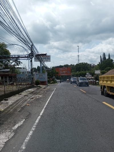 The scene depicts an asphalt road extending into the distance under a sky with scattered clouds. On the left, a dense array of black utility cables spans overhead, connected to poles situated above a metal fence and concrete pedestrian area. Behind these, a white building with a distinctive blue arched roof structure is partially visible. A prominent white billboard displays text including "RESTORAN DEWI LASARI INDAH" in black letters. Further along, a grey sign reads "DPA HOTEL & PRIKULA" followed by additional text. On the right side of the road, a yellow dump truck, laden with granular material, occupies the foreground. Multiple passenger vehicles, including a dark SUV and a black sedan, are traveling away from the viewer in the right lane. An orange billboard displays "Welcome To RAJA M..." with further text. A tall cellular tower is visible in the background, surrounded by lush green trees. No human figures are clearly visible or identifiable.