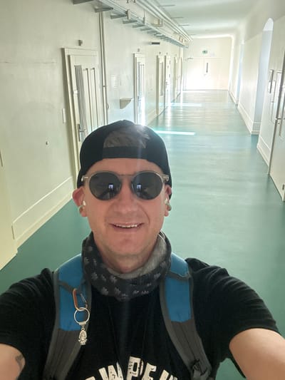 A person with fair skin, wearing a black baseball cap, round sunglasses, a black t-shirt, a grey scarf, and a blue and grey backpack, stands in a long, brightly lit hallway in the city of Bellelay, Switzerland. The hallway has light-colored walls and doors, and a light green floor. There are fluorescent lights on the ceiling, and sunlight streams in from windows at the far end of the hallway. The person is smiling and looking at the camera, taking a selfie. There is a keychain with a small, ornate pendant hanging from the backpack. The overall impression is that of a casual, perhaps tourist, snapshot taken in a clean, institutional-looking environment.