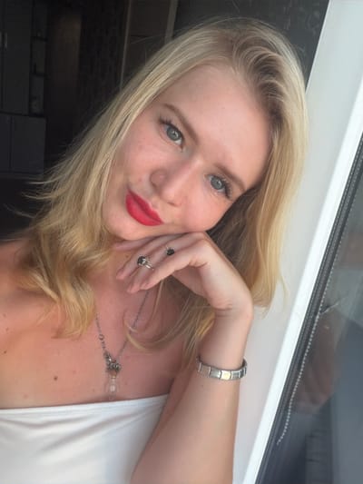 A young Caucasian woman with blonde hair and blue eyes is shown in a close-up, selfie-style shot. She is wearing bright red lipstick and has a white strapless top. Her left hand is resting on her cheek, and she is wearing two rings on her index and middle fingers, along with a silver charm bracelet on her left wrist. A necklace with a glass pendant hangs around her neck. The background is dimly lit with a dark, blurred area to the left and a bright window frame on the right, suggesting an indoor setting. The image does not contain any information that would allow for the identification of its location within the city of Pyt'-Yakh, Russia.
