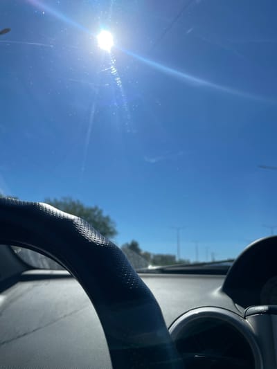 The image shows a view from inside a car, looking out through the windshield on a bright, sunny day. The sun is high in the sky, casting strong glare and lens flares across the scene. The foreground is dominated by a portion of a black steering wheel, textured with a pattern of small dots. To the right, parts of the car's dashboard are visible, including a dark curved surface and the edge of a circular gauge.
Beyond the car's interior, the view through the windshield reveals a clear blue sky. In the distance, trees with green foliage are visible, and a highway or road with lampposts can be seen. The overall impression is that of a car driving on a clear day, likely on a highway or a road with sparse urban development.
The location is specified as Aktau, Kazakhstan. While the image itself does not contain specific visual cues to pinpoint the exact location within Aktau, the context suggests it is a road in or near the city. The presence of the highway and lampposts is consistent with urban or suburban road infrastructure. The sunny weather indicates a typically clear day in the region. There are no people visible in the image, and the focus is on the car's interior and the external environment.
