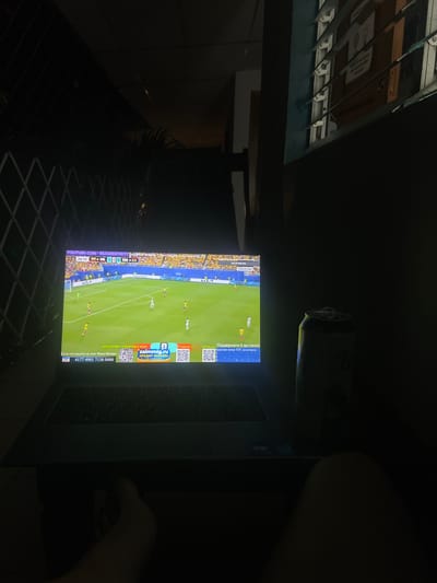 The image shows a laptop displaying a soccer match on a YouTube stream. The stream is overlayed with text and graphics. A can of what appears to be beer is next to the laptop. The scene is outdoors at night. The background includes a wire fence and louvre windows with some posters visible. The overall ambiance suggests a casual, relaxed setting. The location is identified as San Juan del Sur, Nicaragua.