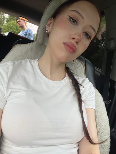 A young woman with long, dark brown hair braided down her back, wearing a white t-shirt, is sitting in the passenger seat of a car. She is looking at the camera with heavy eyeliner and light pink blush on her cheeks. Her lips are slightly parted and appear to be glossed. In the background, a man with short brown hair and glasses is visible in the driver's seat, looking out the window. He is wearing a light blue t-shirt. The interior of the car is visible, with a fluffy white sheepskin seat cover on the passenger side. The setting appears to be outdoors, with trees and greenery visible through the car windows, suggesting a rural or suburban environment. The location is identified as Gavrilovka, Russia.