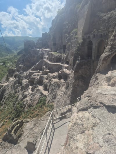 The image depicts the Vardzia cave monastery complex in Georgia, a striking medieval site carved into a cliff face. The primary subject is the monastery itself, with its numerous cave dwellings, churches, and tunnels integrated into the sheer rock. The scene is an outdoor, daytime view showcasing the expansive scale of the complex and its dramatic natural setting.

The image captures a series of stone staircases and pathways, some with metal railings, that snake up and across the cliff, providing access to the various cave levels. In the upper part of the image, larger arched openings suggest significant structures or entrances. Green vegetation grows sparsely on the rocky slopes and in crevices, contrasting with the gray and tan tones of the rock.

Below the cliff, a verdant valley with rolling green hills stretches into the distance, hinting at the remote and mountainous geography of the location. The sky is partly cloudy with bright white cumulus clouds scattered against a clear blue expanse, suggesting fair weather. The lighting indicates it is likely midday or afternoon, with the sun casting shadows on the rock and pathways.

While no people are clearly identifiable as main subjects, the presence of the pathways and railings suggests that visitors or monks would traverse this complex, implying human activity. The overall impression is one of awe at the architectural ingenuity and the historical significance of this ancient monastic site, set against a beautiful natural backdrop. There is no visible text in the image.