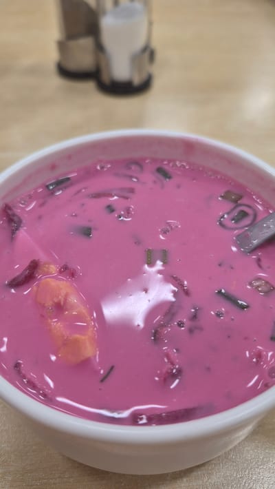 A close-up, eye-level shot shows a bowl of pink soup on a wooden table. The soup appears to be cold, with visible chunks of what look like beets and possibly some herbs. A piece of boiled egg is also discernible in the soup. A spoon is partially visible on the right side of the bowl. In the background, out of focus, are salt and pepper shakers. The setting is likely a restaurant or cafe in Preiļi, Latvia.