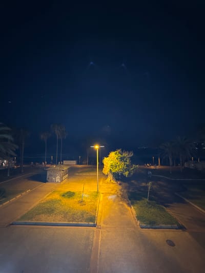 The image shows a street scene at night in the city of Arona, Spain. A brightly lit street lamp illuminates a paved walkway and a grassy area with a small tree. In the background, palm trees line a beach area where several people are visible. A small, graffiti-covered structure sits near the walkway. The sky is dark and clear, with some distant lights visible on the horizon.