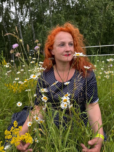 A woman with vibrant red, curly hair and bright red lipstick sits in a field of wildflowers. She is wearing a dark blue dress with white polka dots. Her head is tilted, and she is looking off to the right. Daisies and other small yellow and purple flowers are growing all around her. In the background, there are tall green trees and a white fence. The lighting suggests it is daytime. The woman's hands are visible, one resting in her lap and the other holding a bundle of grass. She appears to be wearing a yellow wristband on her left arm. The scene is set in a natural, outdoor environment, likely a meadow or field.