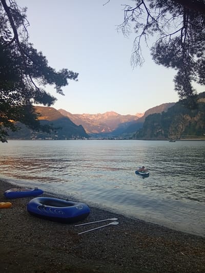 The photograph captures a serene lakeside scene in Hergiswil (NW), Switzerland, at dusk. The foreground is dominated by a pebbled shore where two inflatable blue boats are resting. One boat is closer to the viewer and is dark blue with lighter blue accents and a white circle with "200" inside. The second boat, further back, is also blue. Two white oars lie on the shore between the boats. A bright yellow object, possibly another inflatable, is partially visible behind the closer boat.

The lake stretches out into the midground, its surface rippling gently. In the water, a person is seated in a small, round, grey inflatable, looking towards the shore. Further out on the lake, a sailboat is visible on the right side, moving away from the viewer.

The background features a picturesque landscape of rolling hills and mountains. The sun, low on the horizon, casts a warm, golden light on the peaks, creating long shadows across the slopes. A town with buildings is nestled along the base of the hills on the far side of the lake. The sky above is a pale, clear blue, transitioning into lighter shades near the horizon. Silhouetted branches of trees frame the top of the image, adding a natural, organic border to the composition. The overall atmosphere is peaceful and tranquil, suggesting the end of a warm day.