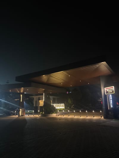 The image shows a nighttime scene outside a building with modern architecture. The building features a wooden-paneled overhang illuminated by embedded lights. In the background, there are illuminated signs. One sign, which appears to be a neon sign, reads "SEMASA" in large, stylized letters. Another sign above it, also illuminated, has text that is partially legible, including "GARAGE" and "FILTER" in smaller print on a dark background with a red border. The ground is paved with brick-like stones, and a row of evenly spaced bollards with upward-facing lights lines a walkway. The overall ambiance suggests a well-lit commercial or public space at night, possibly an entrance to a shopping center or entertainment venue, given the prominent signage. The location is identified as Babakan Madang, Indonesia. There are no people clearly visible in the image. The lighting suggests it is evening or nighttime.