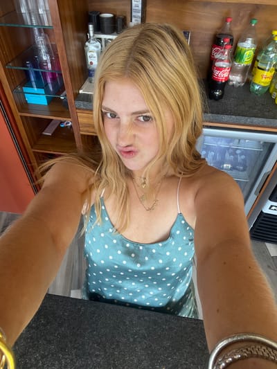 A woman with blonde hair poses for a selfie. She wears a light blue tank top with white polka dots and multiple necklaces. Behind her is a wooden cabinet containing glasses, liquor bottles, and colored boxes. Further back, various soda bottles sit on a counter next to a cooler. The photo appears to be taken from a high angle, showing the woman's arm extended towards the camera, adorned with a thick bracelet.