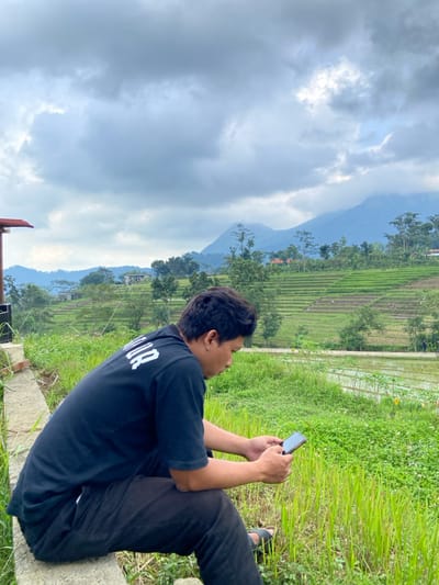 A single adult male is depicted in a rural landscape near Penanggungan, Indonesia. He is seated in profile, facing right, on a light gray concrete ledge in the foreground. He wears a black short-sleeved t-shirt with white lettering on the back, black pants, and dark open-toed sandals. The individual holds a dark-colored smartphone with both hands, looking down at its screen. To his immediate left, a portion of a building with a red roof is visible. The mid-ground reveals extensive green terraced rice paddies, some areas appearing flooded or freshly planted, interspersed with various trees and several small structures featuring reddish roofs. A narrow, light-colored path or road crosses horizontally through the terraced fields. The distant background is dominated by large, hazy blue-green mountains, partially obscured by cloud cover. The sky is uniformly overcast with dense gray clouds, indicating diffused lighting conditions.