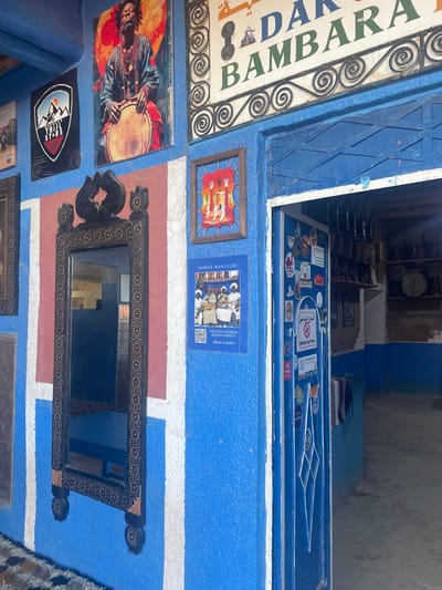 A building facade in blue and white features a sign reading "DAR BAMBARA" and diverse decorations. A colorful Jimi Hendrix image adjoins a mountain range logo. Mirrors with decorative frames and framed art pieces line the wall. A blue poster promotes "HAMAD MANJOUBI KRAMILA MOROCCO." The blue entrance door, adorned with stickers, leads to a dimly lit shop interior.