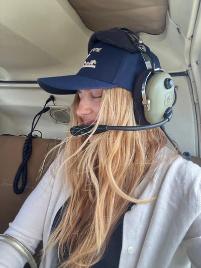 A blonde-haired person wearing a navy blue baseball cap and aviation headset is seated inside an aircraft, presumably on the ground. The person is wearing a white button-up shirt. A speaker or vent is visible next to their face. Part of a brown seat is also visible, along with the aircraft's interior. There is no specific indication of a location within Victoria Falls Municipality, Zimbabwe.