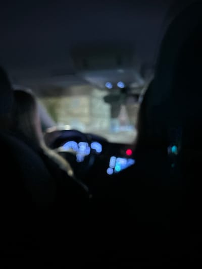 The image is a dark, blurred shot taken from inside a vehicle at night. The car's dashboard and instruments are visible as blue and red lights. Outside the car window, indistinct shapes suggest a building or street scene, illuminated by blurred streetlights. The foreground is dominated by dark shapes, likely parts of the car's interior, obscuring much of the view. The focus is soft, making details difficult to discern.