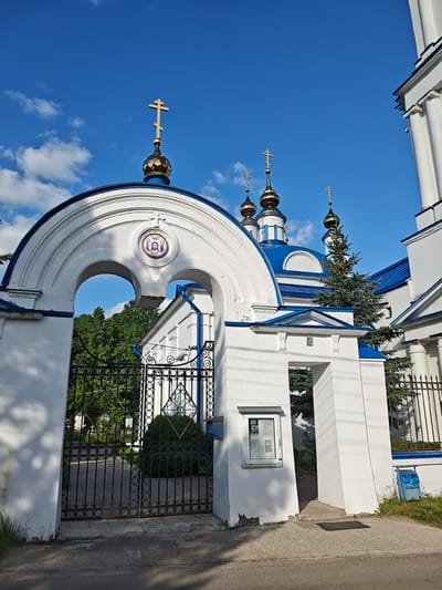 The image shows an Orthodox church in Мамонтово, Russia. The church is white with blue accents on the roof and trim. It features multiple golden domes topped with Orthodox crosses. An arched entranceway with a gate in the foreground leads to the church grounds. A religious icon is visible within the arch. A notice board is attached to one of the gate pillars. Green trees are seen through the gate. The sky is blue with scattered clouds.