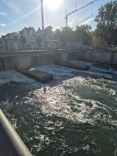 The image depicts a scene at the Isar river in Landshut, Germany, specifically at the dam and weir structure. The sun is bright, casting a glare on the water and creating lens flare in the upper right quadrant.
In the foreground, the dark, turbulent water of the Isar flows rapidly, with white foam and spray generated by the cascading water. A concrete barrier or railing runs along the bottom left edge of the frame.
The central part of the image shows the weir, a man-made structure designed to control water flow. Water is actively spilling over the edge of the weir, creating multiple streams of falling water. In front of the weir, large, rectangular concrete blocks protrude from the water, serving as obstacles or part of the weir's construction.
Beyond the weir, a bridge or walkway is visible. On this walkway, several people are standing and walking, appearing to observe the river or the surrounding area. They are dressed in casual, light-colored clothing.
In the background, a townscape with buildings of varying architectural styles is visible. These buildings have pitched roofs and appear to be typical of European urban architecture. Behind the town, large green trees are present, and further in the distance, construction cranes are prominent against the sky, suggesting ongoing development or infrastructure work in the city. The sky is blue with scattered white clouds.