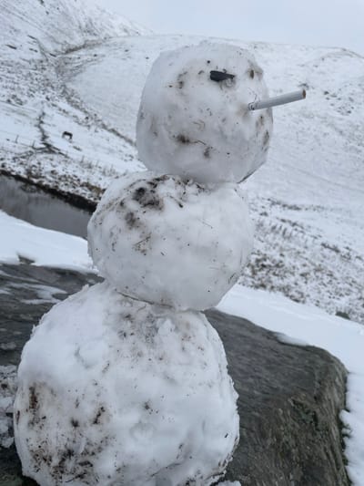 A three-tiered snowman, constructed from snow containing visible soil and plant debris, occupies the foreground, positioned on a large, grey rock formation partially covered in snow. The uppermost snowball, serving as the head, features a single dark, irregular object embedded to represent an eye and a white, unlit cigarette protruding horizontally from the facial area. The two lower snowballs form the body, with the lowest and largest resting directly on the rock. In the middle ground, a narrow body of water reflects the overcast sky, bordered by snow-covered terrain. A faint path or animal trail is visible on the distant, snow-dusted mountain slope, where a dark, equine-like animal stands. The background consists of rolling, snow-covered mountains under a uniformly grey, overcast sky. The scene depicts a winter landscape in the mountainous region of Juta, Georgia.