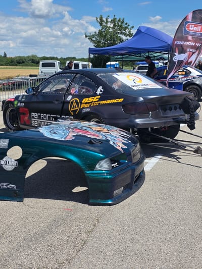 At a sunny outdoor event in Akatsievo, Bulgaria, two modified cars are the primary focus. The foreground features the hood of a dark green vehicle with a colorful graphic of stylized dragon heads and flames. Attached to this hood is a fender with the "NO LIMIT PERFORMANCE PARTS" logo, also featuring a circular graphic. Behind it, a black performance car with racing modifications is parked. Its rear is visible, showcasing wide rear tires, a roll cage, and various sponsor decals, including "OSC Performance" and "DONCHE VHELP." The car has an aerodynamic body kit and is equipped with drag racing tow bars. In the background, a blue canopy tent is set up, with people milling about. Several white vans are parked further back, and a field of dry grass is visible beyond them. The scene is set on an asphalt surface, likely a race track or event venue, under a bright blue sky with scattered clouds.