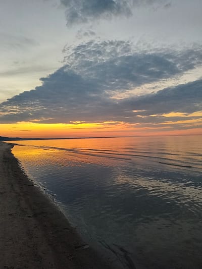 The image depicts a coastal scene at sunset in Apšuciems, Latvia. A dark sand beach occupies the left foreground, showing faint footprints. The Baltic Sea stretches out to the right, with gentle waves lapping at the shore. The water reflects the vibrant orange and yellow hues of the sunset, creating a shimmering effect. In the distance, a dark, silhouetted treeline marks the horizon. The sky above is a canvas of dramatic clouds, with dark gray formations contrasting against lighter gray and pale yellow tones near the horizon. The overall atmosphere is tranquil and serene, characteristic of a coastal sunset.