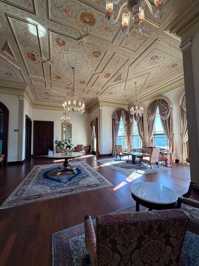This is an interior shot of a grand, ornate room in the Beylerbeyi Palace in Üsküdar, Turkey. The room is decorated in a lavish style with a highly detailed, painted ceiling featuring floral motifs and geometric patterns. Two crystal chandeliers hang from the ceiling, casting a warm light. The floor is polished dark wood, and several large, intricately patterned Persian rugs are laid out.
In the center of the room, a large round table with a marble top stands on a decorative wooden base, adorned with a vase of white flowers. To the right, a seating area features two ornate chairs and a small table, positioned in front of tall, arched windows draped with heavy, golden curtains. Sunlight streams through these windows, illuminating parts of the room and casting dramatic shadows.
Further into the room, another seating area is visible with armchairs and sofas, suggesting a formal reception or drawing-room. The walls are painted in a pale color, accentuating the opulence of the ceiling and furnishings. Wooden doors are visible on the left side of the room. The overall impression is one of historical grandeur and luxurious elegance, typical of Ottoman palace architecture. There are no people present in the image.