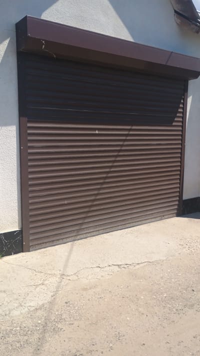 The image shows a closed brown roller shutter door on the side of a building in Mitoc, Moldova. The building's wall is textured white. The ground in front of the door is concrete with cracks and some gravel. A shadow from an unseen object falls across the door and the ground. The scene appears to be outdoors, possibly in a residential or commercial area. There are no people or animals visible in the image.