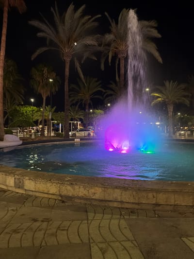The image depicts a nighttime scene at a tropical resort, likely Sant Antoni de Portmany, Spain. The primary focus is a brightly lit fountain in a pool, illuminated with vibrant purple and green lights, creating a magical and inviting atmosphere. The water from the fountain sprays upwards and outwards, catching the colorful light and adding a dynamic element to the scene.

Surrounding the pool are several palm trees, their fronds illuminated by the ambient light and the distant glow of buildings. Their silhouettes against the dark night sky and the fountain's glow suggest a warm, balmy evening. The pool itself is edged by a textured stone wall, adding a natural and robust feel to the setting.

In the background, glimpses of resort buildings and lighting suggest that this is a public area, possibly a hotel or entertainment complex. The lighting and overall ambiance evoke a sense of relaxation and enjoyment, typical of a vacation destination. The time of day is clearly night, given the darkness of the sky and the artificial lighting. No people are clearly visible, but the setting implies human presence and activity in the broader resort. There is no visible text in the image. The overall impression is one of a lively and picturesque resort setting at night.