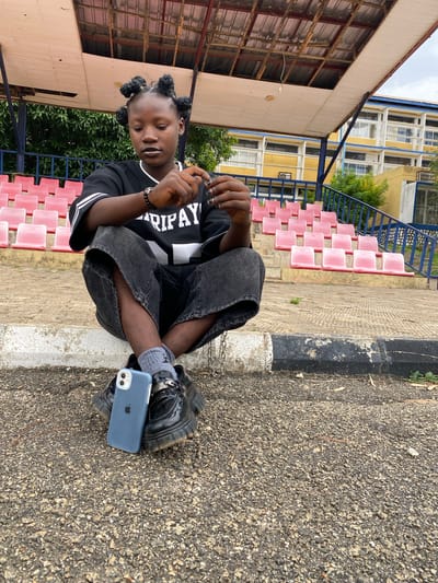 A young Black person with braided hair styled in four buns sits on a concrete step in front of a row of pink stadium seats. They are wearing a black t-shirt with white lettering and trim, dark baggy pants, grey socks, and black chunky shoes. A light blue iPhone is placed on the ground in front of their feet. The background shows a yellow building with blue trim and a covered structure with red seating. The scene appears to be an outdoor stadium or sports facility, possibly in Zawan, Nigeria.