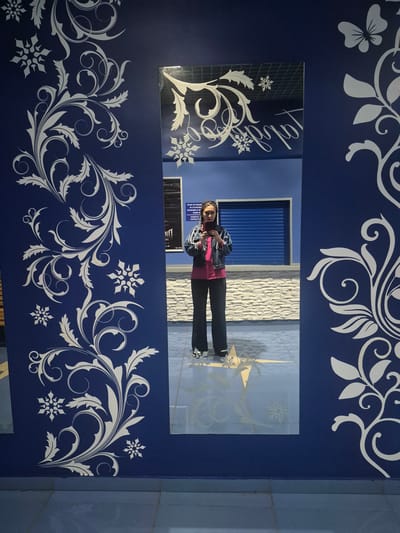 A full-body, selfie-style photograph captures a woman standing in front of a dark blue wall adorned with white floral and snowflake decals. The woman is wearing a pink shirt, a dark denim jacket, and dark pants. She is holding a smartphone in her right hand, taking the picture. A large mirror is present, reflecting the woman and the wall behind her. The mirror also shows a glimpse of a blue roller shutter door and a stone-tiled wall in the background, suggesting an indoor environment. The floor is tiled in a light blue hue with a faint starburst pattern visible. The overall scene is indoors, possibly in a public space or a shop, with decorative elements on the wall. There is no visible indication of the city of Кварса, Russia within the image itself.