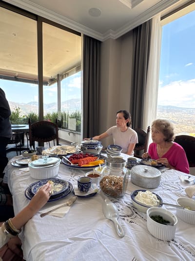 A breakfast scene unfolds indoors. A woman with short brown hair stands at a table covered with a white lace tablecloth, reaching towards a blue teapot and a platter of cut fruit. To her right, an older woman with short grey hair sits and eats. Various serving dishes containing toppings such as granola, cream, and green herbs sit around the table. Outside the window is a view of Santiago Yancuitlalpan, Mexico.