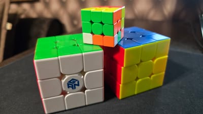 Three Rubik's Cubes are arranged on a black surface, possibly a desk mat. The cube in the foreground left is predominantly white with green and red sides visible. It features a blue logo with the letters "GAN". To the right and slightly behind is another cube, primarily yellow with red and blue sides. A smaller cube sits on top of the yellow and red cube, showing green, orange, and yellow faces. The background is blurred, revealing a dark, textured surface that appears to be upholstery, potentially from a chair or sofa. The lighting casts shadows on the black surface. There are no people or animals present. The scene is an indoor, still-life arrangement of puzzle toys.