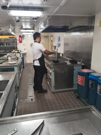 A man in a white t-shirt, black apron, and black pants is standing in a ship's galley, a commercial kitchen. He is positioned on the right side of the image, facing away from the viewer and towards a large stainless steel stove. He appears to be holding a utensil, possibly stirring a pot on the stove. The galley environment is industrial, with stainless steel countertops, shelves, and appliances. There are various kitchen items visible, including pots, pans, and utensils. On the left side of the image, there are large stainless steel sinks and a rack of eggs. In the background, there are blue waste bins and signage indicating waste classification. The flooring is a grid pattern of dark tiles. The lighting is fluorescent. The scene appears to be in a functional, operational kitchen. The image does not provide enough information to definitively place the location in Velsen-Noord, Netherlands.