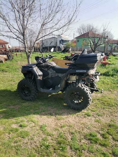 The image shows a CFMOTO ATV parked on a grassy lawn in an outdoor setting. In the background are a tree with bare branches, a white truck, a green house, and other farm equipment. The ATV is a dark color with camouflage patterns, has black wheels, and a black storage box mounted on the back. Based on the geotag metadata the location is Paisievo, Bulgaria.