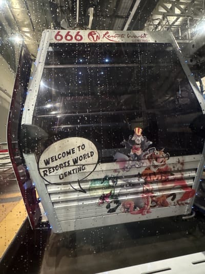 The image shows a glass-enclosed structure labeled "Resorts World Genting" with the number "666" on one side. A sign says "Welcome to Resorts World Genting." Character cartoon imagery is also present. The glass is covered in water droplets, suggesting rain or condensation. The setting is likely Genting Highlands, Malaysia, based on the labeling. The structure appears to be a transportation unit, potentially a cable car or enclosed vehicle, seen through a wet glass surface. The surrounding environment is obscured but shows metallic framework and indistinct shapes.