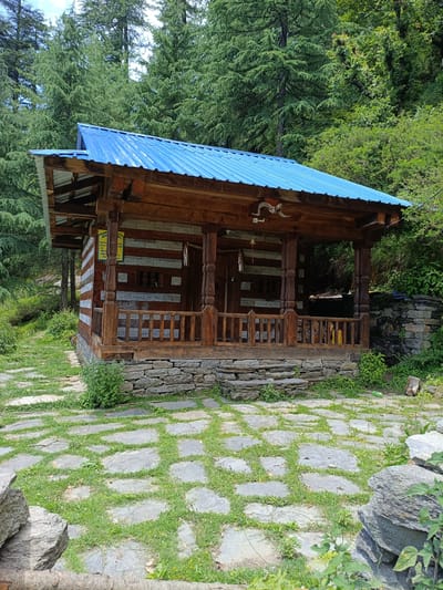 The image depicts a small, wooden building with a blue metal roof, nestled within a dense forest. The building has a covered porch with wooden pillars and a railing. The walls appear to be constructed of horizontal wooden logs alternating with stone. A stone pathway, with grass growing between the stones, leads up to the entrance of the building. The surrounding environment is lush and green, with tall trees visible behind the structure. There are no people present in the image, and the scene appears to be tranquil and natural.