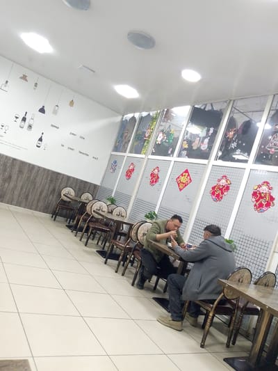 The image shows the interior of a restaurant with two men seated at tables. One man in a green jacket is looking at his phone while holding cutlery. The other man in a gray hooded jacket is also looking at his phone. The restaurant has a tiled floor and walls decorated with images of wine bottles, glasses, and pendant lights. The windows are adorned with festive decorations, including images of Santa Claus, Christmas trees, and Chinese New Year ornaments. The overall ambiance suggests a casual dining setting. The image is not located in Magadan, Russia; it is a generic restaurant interior.
