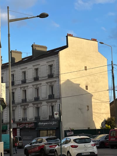 The image depicts a street scene in Saint-Brice-sous-Forêt, France. It features two buildings, one with a plain facade and the other with ornate architectural details including wrought iron window ornamentation. A lamppost is visible in the foreground. Street signs for "SUPERETTE DE LA GARE", "ST BRICE FORET" are noticeable. Signs for "PIZZA" and "POULET" and the storefront name "Boulangerie de la Gare" suggest the presence of food businesses. A row of parked cars including a white "CLIO" is along the side of the street. A sign indicates a speed limit of 30 km/h. A man is seen in the background. Power lines cross the upper part of the image.