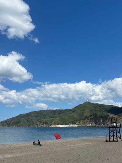 The image shows a beach scene in M'Diq, Morocco. The view is rotated 90 degrees clockwise. The sky is blue with scattered white clouds. A mountain covered with green vegetation runs along the center of the image. The beach is sandy and meets a body of water, likely the Mediterranean Sea.
A person is lying on the beach in the lower right corner, possibly sunbathing. A red umbrella is planted nearby. A black metal structure is near the water’s edge further up the beach. White buildings are visible along the coastline.