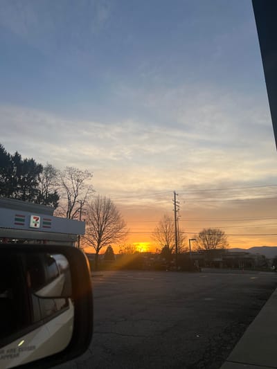 The image captures a sunset scene in what appears to be a commercial area. A 7-Eleven store is visible on the left side, identifiable by its logo. The setting sun casts a bright orange glow on the horizon, silhouetting trees and utility poles. A building that might have a Chili's restaurant is also discernible. A car side mirror is visible in the lower right corner, with the reflection of other vehicles. The photograph seems to be taken from inside a car, and the environment suggests an urban or suburban setting in the City of Newburgh, United States of America.
