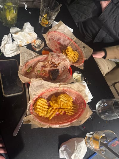 This image captures a close-up, top-down view of a dark table laden with post-meal items in what appears to be a casual dining establishment in Saint-André-lez-Lille, France. The scene indicates a recently concluded or ongoing meal for multiple individuals.
Three red plastic baskets, each lined with parchment paper, are present. The topmost basket contains a small quantity of waffle fries. The middle basket holds remnants of a meal, including a dark, charred piece of food (possibly meat), sauce smears, and green garnishes, suggesting it was largely consumed. The bottom-most basket contains a substantial portion of waffle fries. Several small, clear plastic containers with various sauces or dips, one white and creamy, another orange/red, are also on the table.
Drinkware includes a clear glass in the upper left filled with a beverage, lime wedges, and ice. To its right, an empty, textured glass contains orange or lemon peels. A clear wine glass lies on its side, empty, near the bottom right, and another glass, partially visible, stands upright in the far bottom right.
Other objects scattered across the table include several crumpled white paper napkins. A dark smartphone with a visible screen protector is positioned face down on the left side of the table, and a silver-colored utensil (fork or knife) lies beside it.
Partial views of at least two individuals are discernible. In the upper right, a person's torso and arm are visible, clad in a dark, quilted jacket. Below them, another individual's arm and lower torso are seen, wearing a light tan or beige coat. A hand is also partially visible in the bottom left corner of the frame. The overall impression is a casually disordered table following a social dining experience.