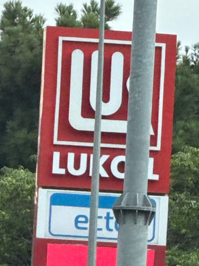 A red and white sign with the Lukoil logo and the word "LUKOIL" is visible. Below it, a white rectangle with a blue border contains the letter "e". A pink rectangle is at the bottom of the sign. The sign is located in Baku, Azerbaijan. A metal pole is in front of the sign, obscuring part of it. Trees are visible in the background.