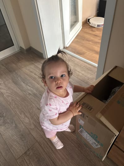 A baby wearing a pink onesie with a bow pattern and striped socks is holding onto a cardboard box. The baby has dark curly hair pulled up and is looking at the camera with wide blue eyes and an open mouth. The setting appears to be indoors, with a wooden floor and a glimpse of white cabinetry or furniture. In the background, a white object that resembles a robot vacuum cleaner is visible on the wooden floor. The lighting suggests it could be daytime. There is text on the cardboard box, including a barcode and what appears to be product labeling. The overall mood is playful and curious, as the baby interacts with the box.