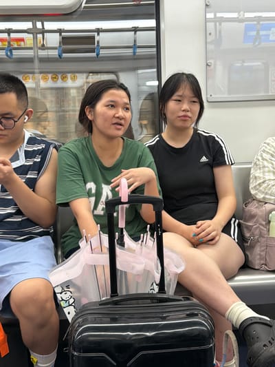 This media file captures three people seated inside a public transport vehicle, possibly a train or bus, given the handrails and window reflections suggestive of a travel setting. The scene appears to be indoors and illuminated by artificial light, indicating it could be any time of day.

The individuals are a young woman in the foreground wearing a green t-shirt, a young woman in the background wearing a black t-shirt with white stripes, and a person whose face is partially visible in the lower foreground wearing glasses and a striped shirt. The young woman in the black t-shirt is leaning back, her legs extended. The woman in the green t-shirt is looking to her left, with her hands resting on a pink and black object. The person in the lower foreground is looking down. The overall mood seems relaxed, with the subjects appearing to be traveling or waiting.

Notable details include a black suitcase next to the woman in the black t-shirt, and a pink garment draped over a black rack, possibly a stroller or cart. The woman in the black t-shirt has blue nail polish. The woman in the green t-shirt has a light-colored garment with text that is partially obscured but appears to include the word "Cha" and some other characters. The background shows the interior of the vehicle with more handrails and reflective surfaces. The location context is provided as Tiaodeng, China.