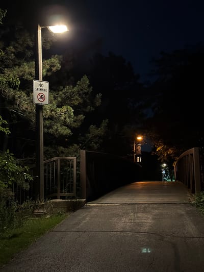 A pathway illuminated by a streetlamp at night, leading to a bridge. A "No Swimming" sign is visible on a pole to the left of the pathway. The pathway appears to be made of asphalt and leads over the bridge. Trees and foliage surround the pathway and bridge. The sky is dark, with a single star visible. The scene is set in the city of Vernon Hills, United States of America.