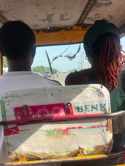 The image shows the interior of a vehicle, likely a tuk-tuk, viewed from behind the passengers. Two people are seated in the front, facing forward. The person on the left has dark, short hair and is wearing a white t-shirt. The person on the right wears a dark green headwrap and has a large amount of reddish-orange dreadlocks. In front of them, a white box with colorful markings is visible. The box has the word "BENK" and some circular red markings painted on it. Through the cracked windshield, a street scene is visible with other vehicles and greenery. The ceiling of the vehicle is decorated with a pattern of stars and cartoon pandas. The overall scene suggests a candid moment of travel within a city, possibly in Galadimawa, Nigeria, given the context provided.