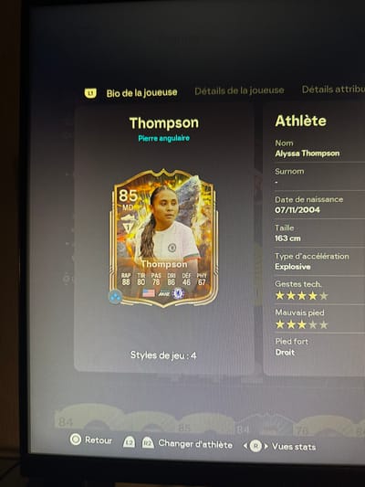 A digital screen displays a video game interface, likely a football simulation, featuring a player profile in French. The primary subject is a golden-bordered player card for "Thompson" with an overall rating of "85 MD" (Milieu Droit/Right Midfielder). The card depicts a female athlete, Alyssa Thompson, from the chest up, with dark braided hair, wearing a white and blue collared shirt. Her in-game statistics are visible on the card: RAP 88, TIR 80, PAS 78, DRI 86, DEF 46, PHY 67. Below these, an American flag, EA Sports logo, and Chelsea FC logo are present.

To the right of the card, an "Athlète" (Athlete) information panel details "Nom: Alyssa Thompson," "Date de naissance: 07/11/2004," "Taille: 163 cm," "Type d'accélération: Explosive," "Gestes tech.: ★★★★★" (5 stars), "Mauvais pied: ★★★★★" (5 stars), and "Pied fort: Droit" (Right foot). A menu bar at the top includes "Bio de la joueuse," "Détails de la joueuse," and "Détails attributs." At the bottom, game control prompts are visible: "Retour" (Return) with a circle button, "Changer d'athlète" (Change Athlete) with L2/R2, and "Vues stats" (View Stats) with R. The screen itself shows minor reflections of a dimly lit room.

The image contents do not provide any visual information to determine its physical location in the city of Kaysersberg Vignoble, France.