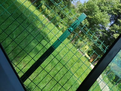 The image is an overhead view, looking down through a green chain-link fence onto a grassy area with trees. The fence has a dark horizontal bar casting a shadow across the scene. In the foreground, a green metal bracket is attached to the fence. The background shows a bright green lawn, with patches of darker green indicating trees and bushes. Some yellow flowers are visible in the grass. In the distance, there appear to be some wooden structures, possibly benches or picnic tables, and what looks like a yellow swing set. The bright sunlight suggests it is a clear day, likely midday. The overall impression is of an outdoor recreational space, possibly a park or a backyard. No people are visible in the image, and there is no visible text. The location context provided is Madan, Bulgaria.